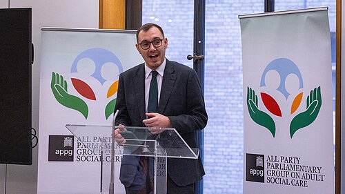 Tom Gordon stood at a podium with signs reading 'All Party Parliamentary Group on Adult Social Care'