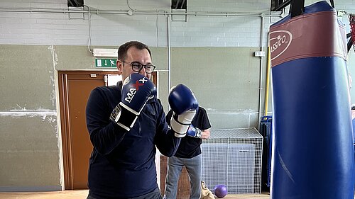 Tom Gordon at a boxing gym with arms up, boxing gloves on and looking at a punchbag